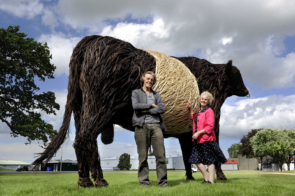 FREE PICTURE: Royal Highland Showcase Wicker Beltie and Tunnel Mural, Edinburgh, 14/06/2021:
Royal Highland Showcase Celebrates Dumfries and Galloway with One-Tonne Willow Bull:
A one-tonne wicker Beltie bull, hand sculpted in Dumfries and Galloway, has arrived in Edinburgh where it will be a centrepiece of this week’s Royal Highland Showcase - at the Royal Highland Showground on the fringes of Edinburgh 
Nearby, the famous underpass where the finest livestock are led in and out of the Ingliston showground arena, has been painted with a magnificent mural depicting farm animals, horses, wildlife and scenes from south-west Scotland.
 The Royal Highland Showcase runs online from today (Monday) 14th to (Sunday) 20th June (2021) and replaces the annual Royal Highland Show which cannot take place due to COVID-19 restrictions - see: https://www.royalhighlandshow.org 
 Each year RHASS invites a different region to act as “host”, allowing it to highlight the best of its rural economy. This year it’s the turn of Dumfries and Galloway which prides itself on its thriving creative community.
  Pictured with the beltie is artist Trevor Leat (correct), whose sculptures are familiar from Edinburgh’s Hogmanay celebrations, the Wickerman music festival and a variety of National Trust for Scotland properties. And artworks project leader Cathy Agnew (correct).
For more information see accompanying press release or contact  Matthew Shelley at Matthew@ScottishFestivalsPR.Org - 07786 704 299. 
 Photography for Royal Highland Society of Scotland (RHASS) from: Colin Hattersley Photography - www.colinhattersley.com - cphattersley@gmail.com - 07974 957 388.