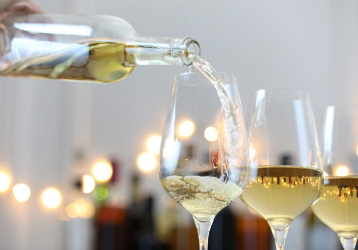 Pouring white wine into glass against blurred background with festive lights, closeup. Bokeh effect Credit: Adobe Stock